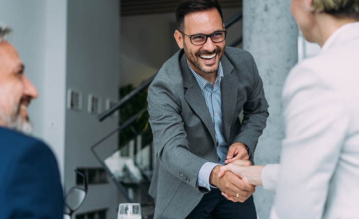 Two people shaking hands in a professional setting. One wears a grey blazer with a blue shirt, and the other wears a white outfit. A third person is partially visible on the left. Background includes a staircase and framed pictures. Faces are blurred for privacy.
